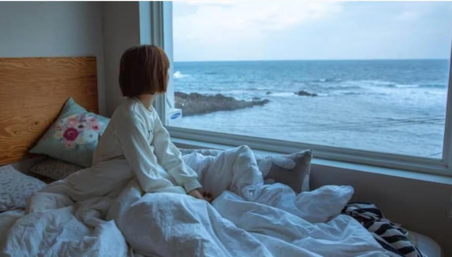 a little girl sitting on a bed looking out a window at Sometimes Jeju in Jeju