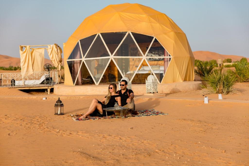 two people sitting in the sand in front of a tent at Sunrise Sahara Camp in Merzouga