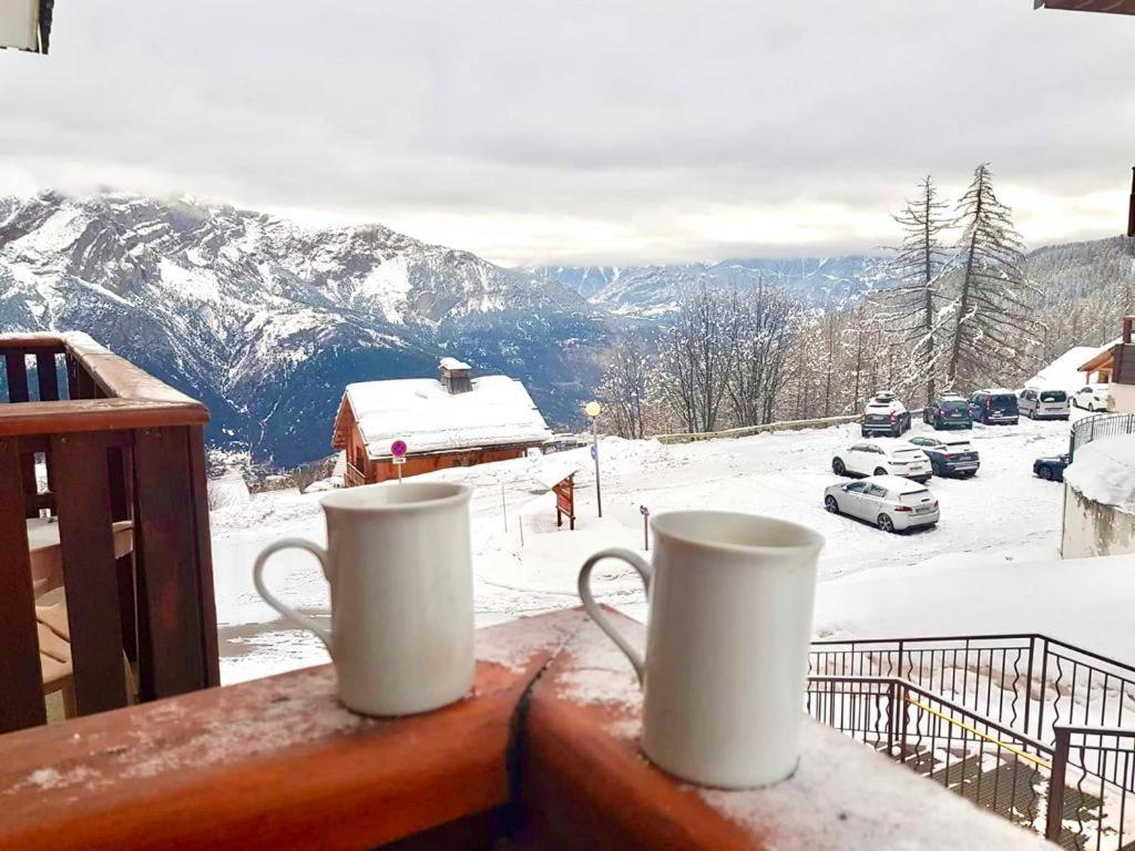 two coffee mugs sitting on a balcony with a snow covered mountain at Le studio 1800 aux pieds des pistes in Puy-Saint-Vincent