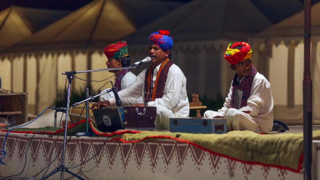 Trzech mężczyzn w turbanach siedzących na scenie z mikrofonem w obiekcie Charisma Desert Camping w mieście Jaisalmer