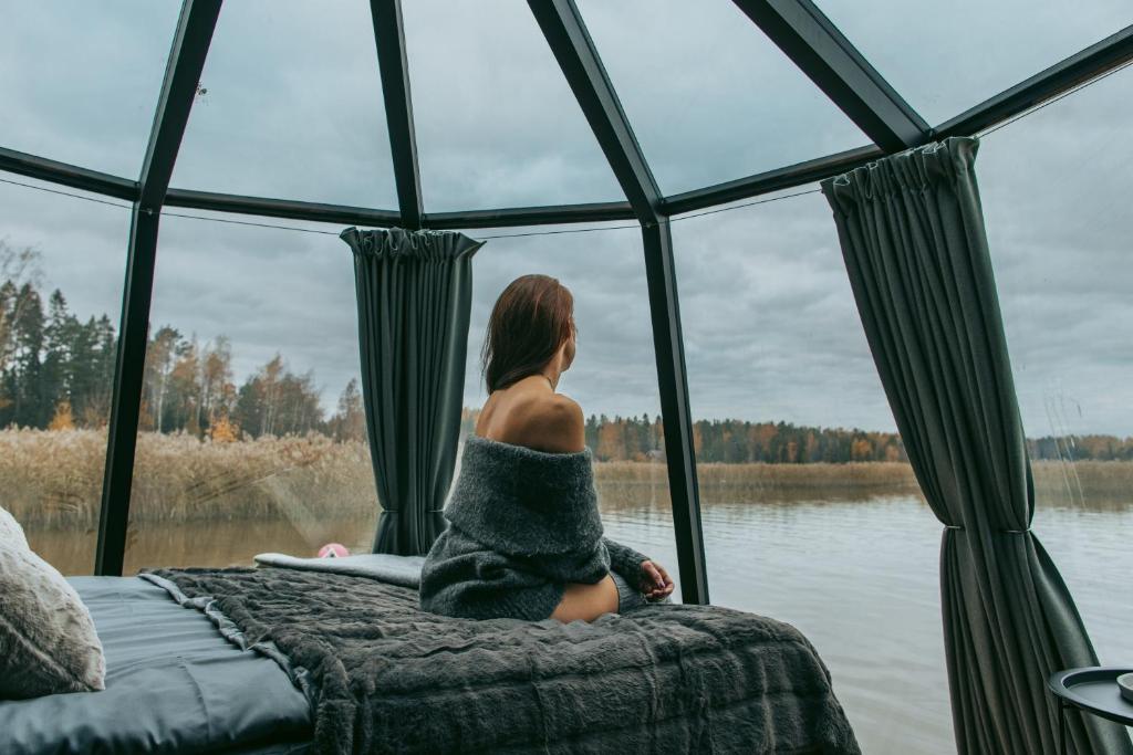 uma mulher sentada numa cama com vista para um lago em Aurora Igloo Porvoo em Porvoo