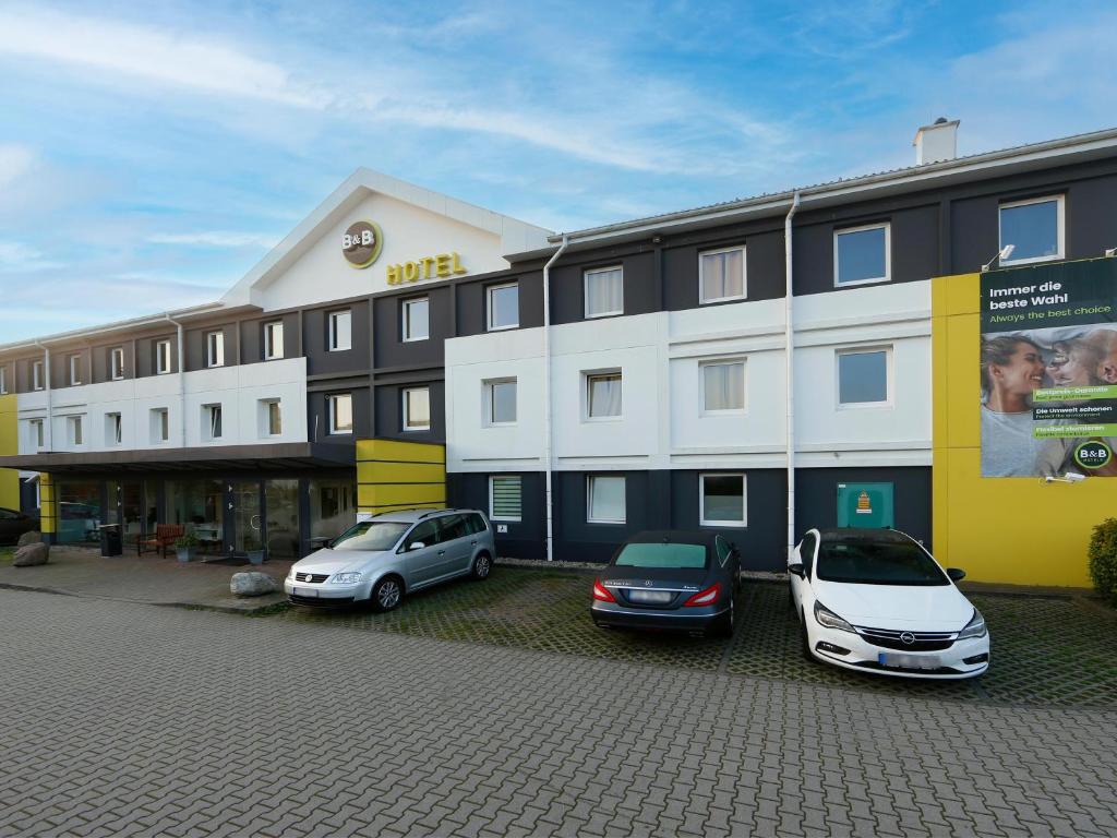 dwa samochody zaparkowane przed dużym budynkiem w obiekcie B&B Hotel Berlin-Süd Genshagen w mieście Ludwigsfelde
