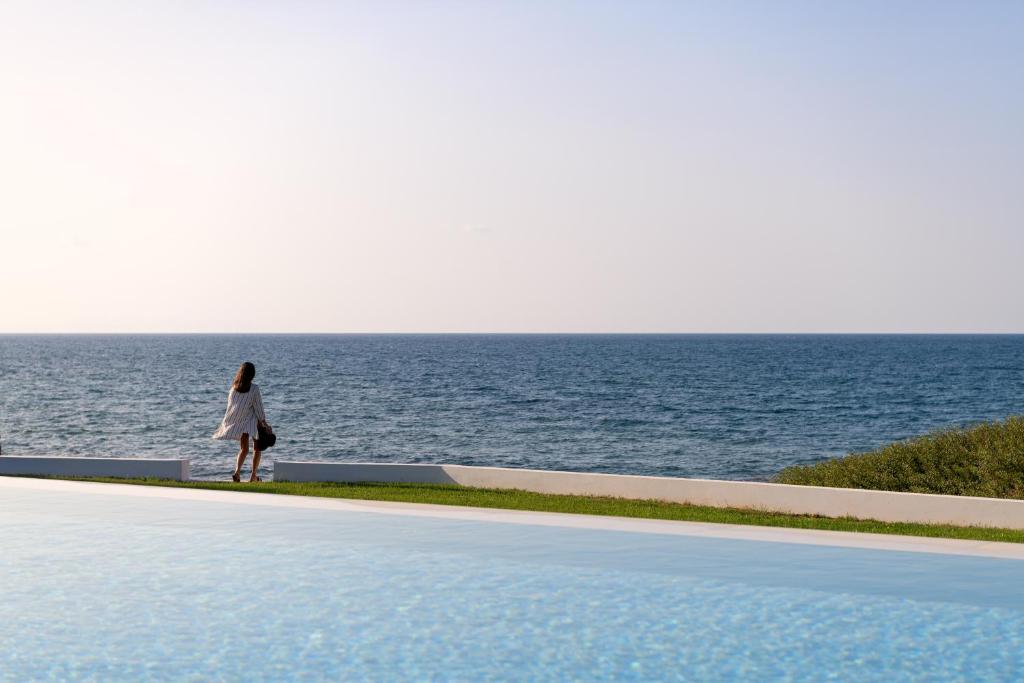 une femme debout à côté d'une piscine à côté de l'océan dans l'établissement Asea Villa, Spectacular right-on-the-beach Retreat, By ThinkVilla, à Skaleta