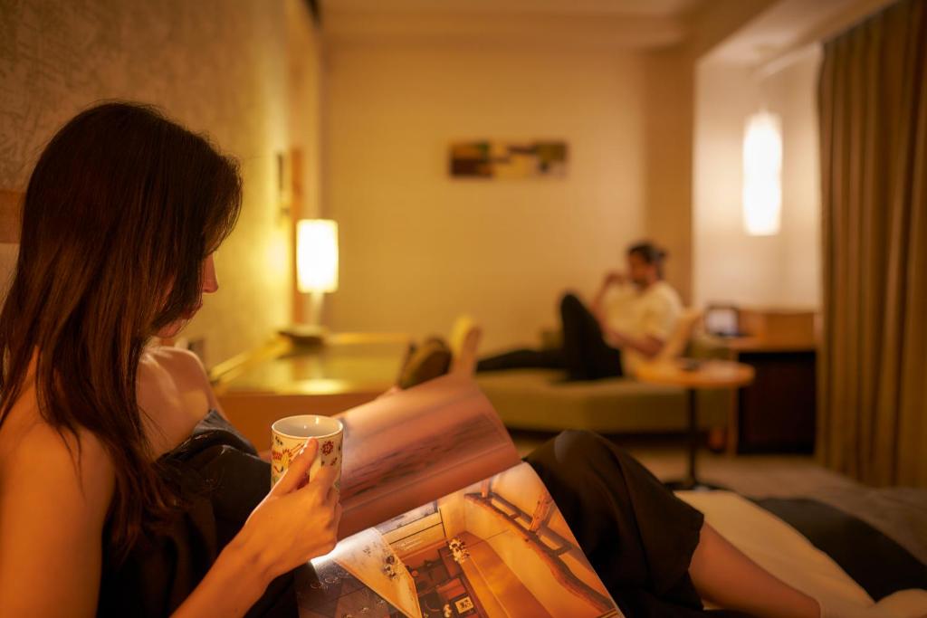 a woman sitting on a couch reading a book at Hotel Resol Trinity Kanazawa in Kanazawa