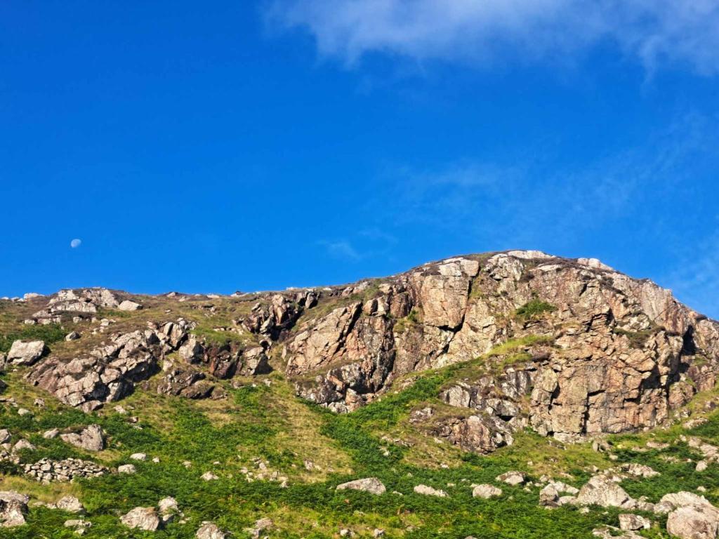 a rocky mountain with green grass and a blue sky at Cliff Cottage in Valtos