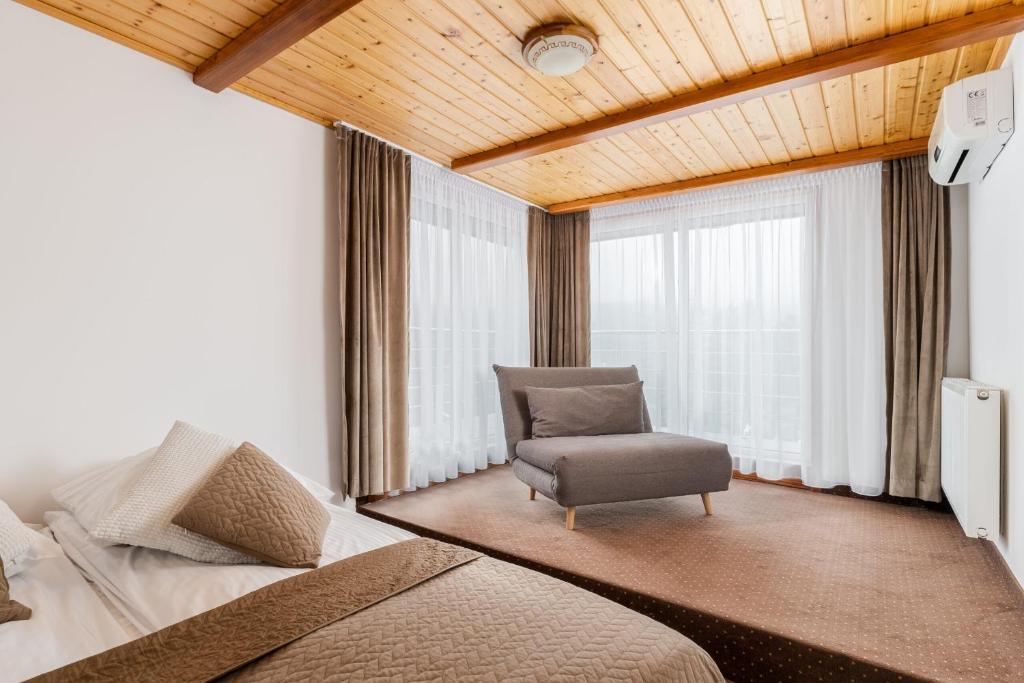 Spacious hotel bedroom corner with wooden ceiling, chaise lounge by large draped windows, neutral bedding.