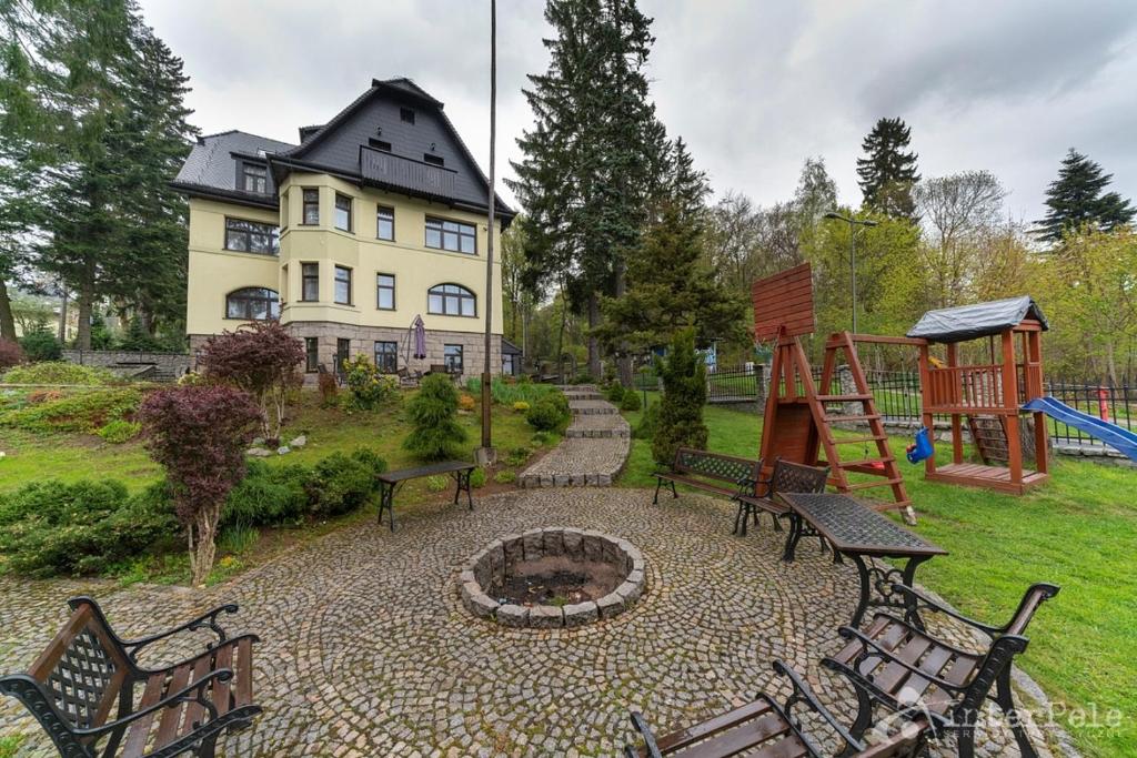 Large yellow villa with cobblestone patio, central firepit, benches and wooden playground, overcast.