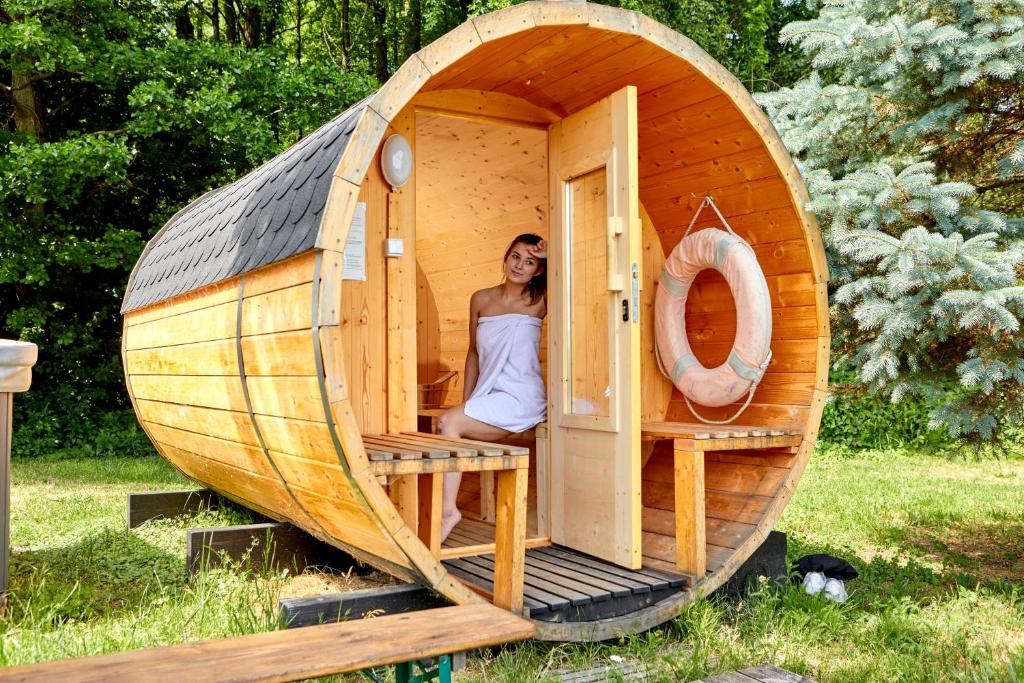 Woman wrapped in towel sitting inside wooden barrel sauna set outdoors near trees.
