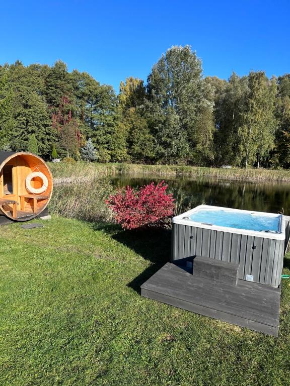 Outdoor grassy lakeside scene with bubbling hot tub, wooden barrel sauna, and surrounding trees
