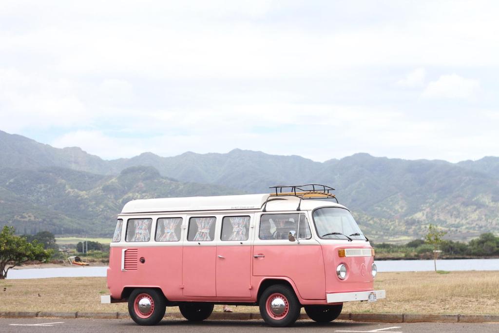 Una furgoneta rosa y blanca conduciendo por una carretera en Hawaii Surf Campers, en Wahiawa