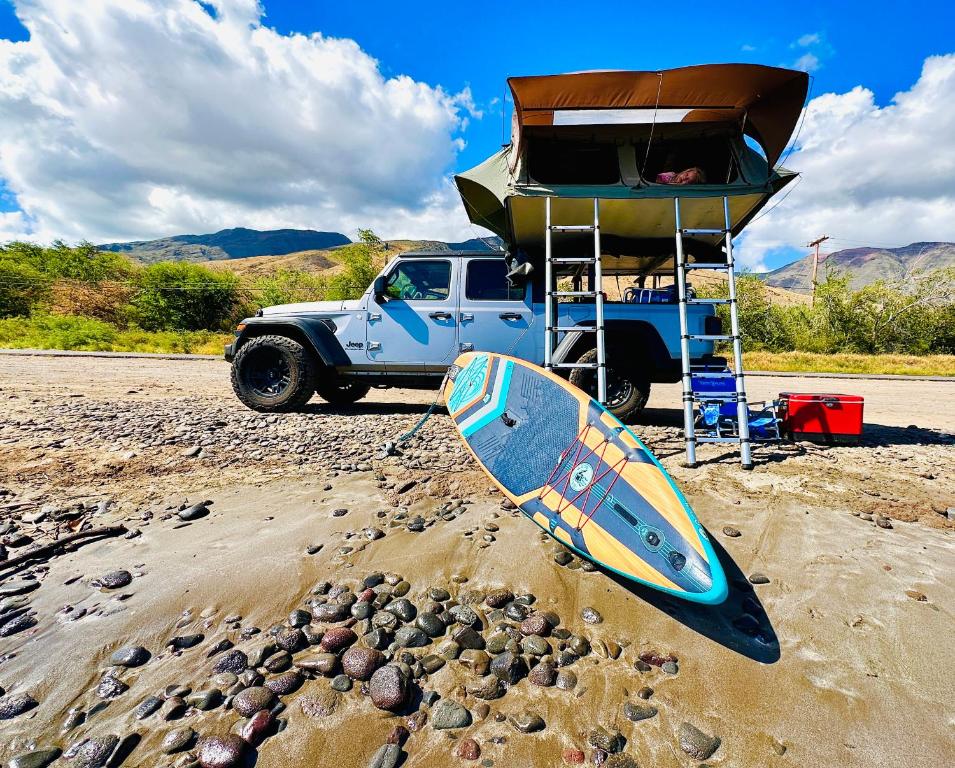 a surfboard sitting on the beach next to a truck at Explore Maui's diverse campgrounds and uncover the island's beauty from fresh perspectives every day as you journey with Aloha Glamp's great jeep equipped with a rooftop tent in Paia