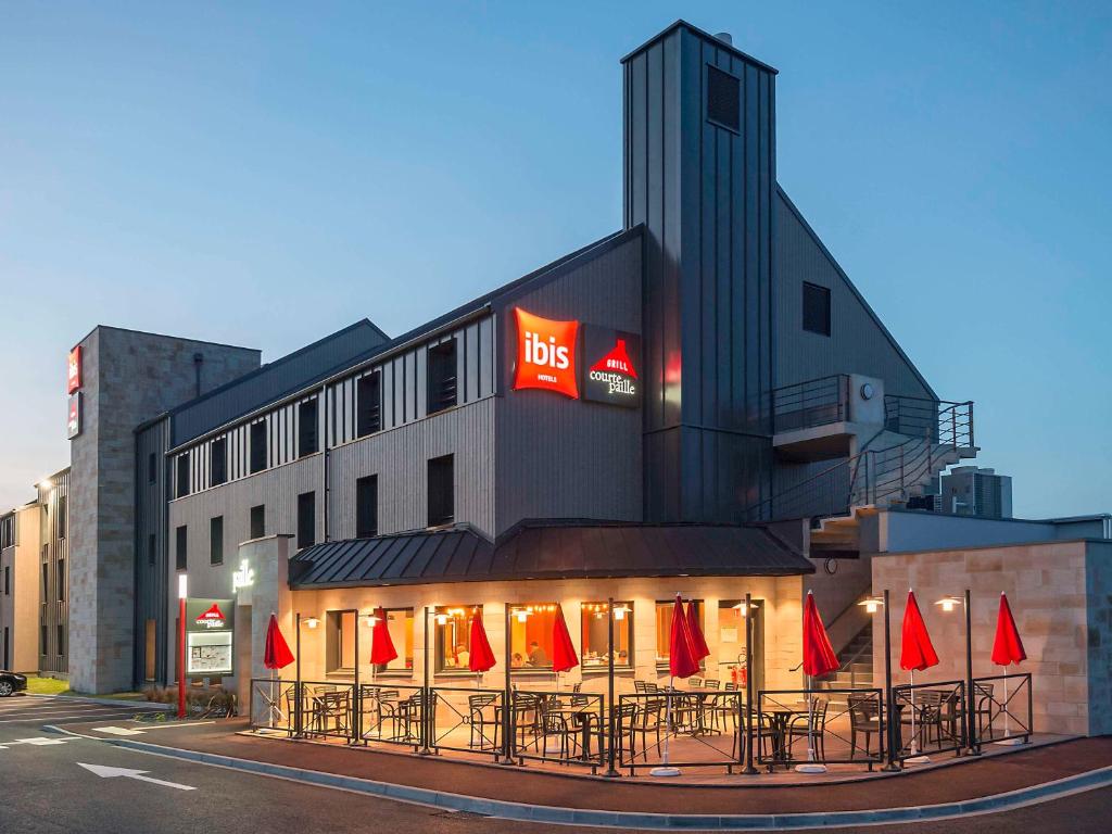 un edificio con mesas y sombrillas rojas delante de él en ibis Pontorson Baie Du Mont Saint Michel en Saint-Georges-de-Gréhaigne