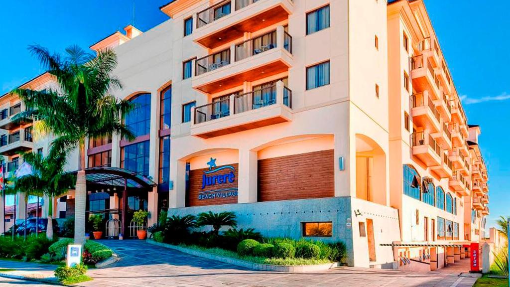 a large building with a palm tree in front of it at Jurerê Beach Village Studios Para Temporada in Florianópolis