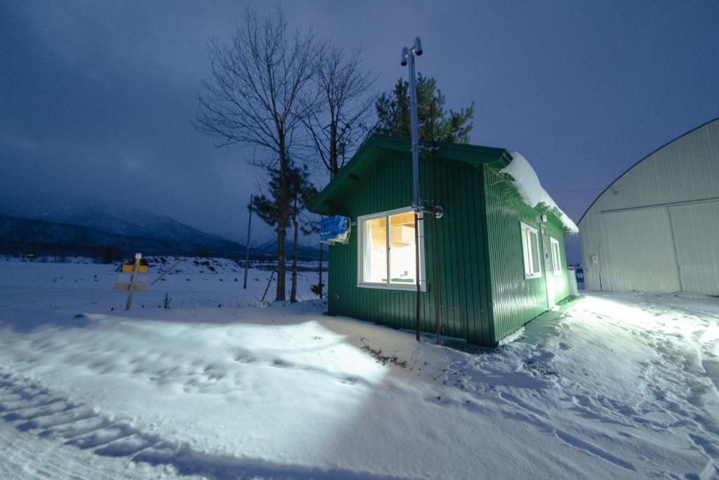 un pequeño edificio verde en la nieve junto a un edificio en Furano Log House Farm Resort, en Furano