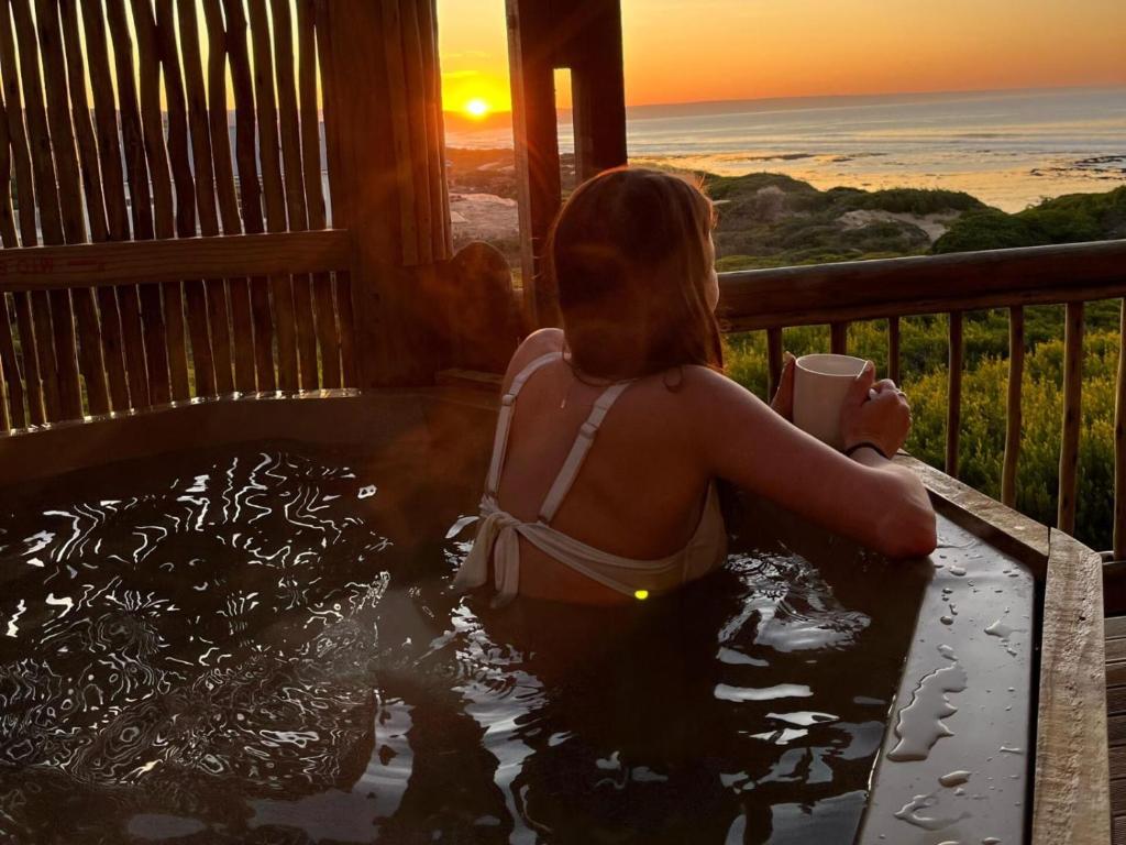 una mujer sentada en un jacuzzi viendo la puesta de sol en Witsand Whalecliff accommodation en Witsand