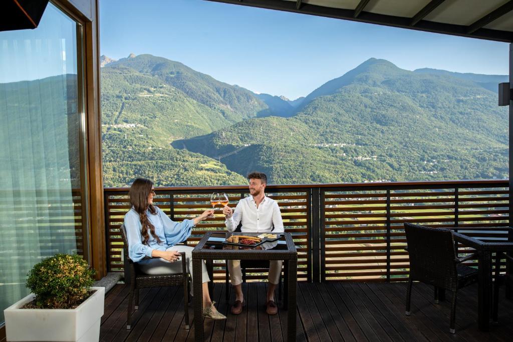 two people sitting at a table on a balcony with mountains at Wine Hotel Retici Balzi in Poggiridenti