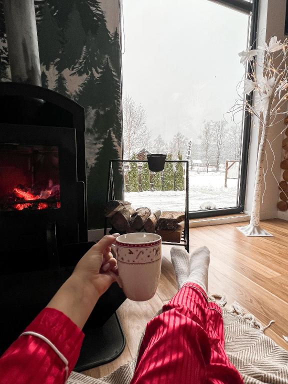 a person holding a cup of coffee in front of a fireplace at Bieszczadzkie BOHO stodoły z jacuzzi i sauna na wyłączność in Myczkowce