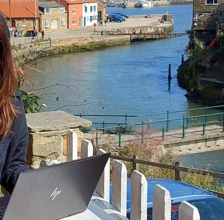 uma mulher sentada numa mesa com um laptop em frente a um rio em Sea Haven holiday cottage at Staithes em Staithes