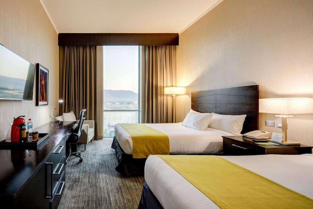 a hotel room with two beds and a desk and a window at La Quinta by Wyndham Santiago Aeropuerto in Santiago