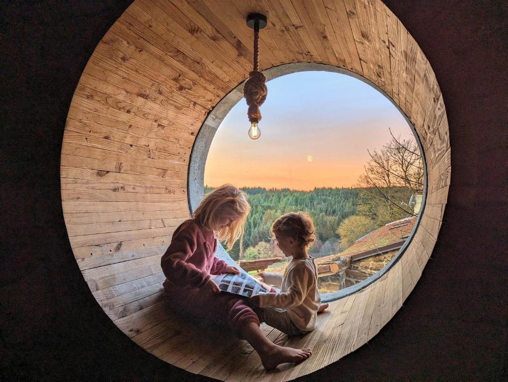 two girls sitting in a round window looking at the sunset at L'œil des Bois - Gîte Familial - Vue panoramique in Lapte