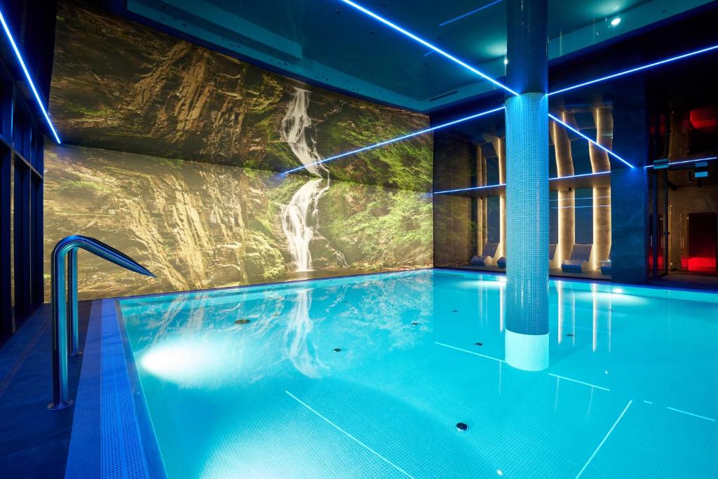 Blue-lit indoor pool with waterfall mural backdrop and glowing linear ceiling lights, modern spa.
