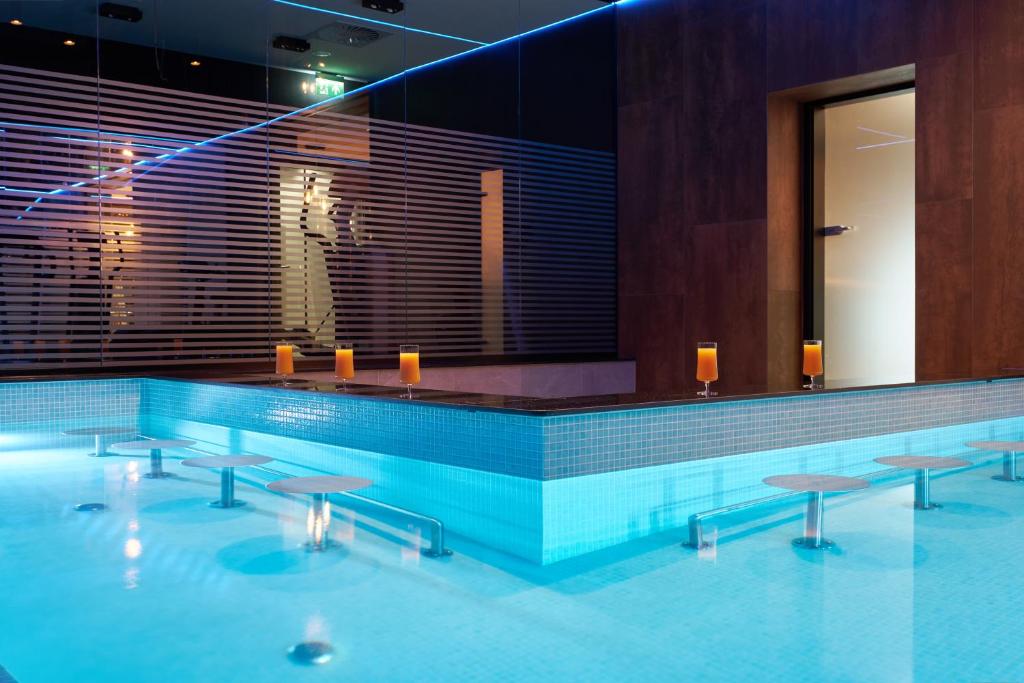 Submerged bar stools and poolside counter with three orange drinks, cool blue lighting.