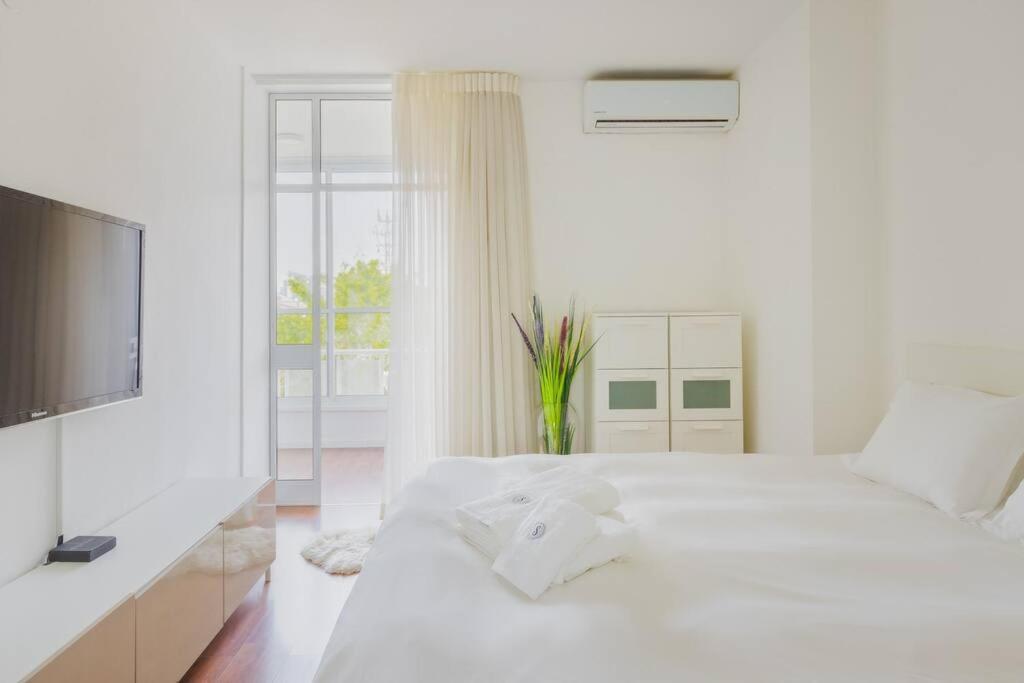 a white bedroom with a white bed and a television at Tiran Square Haven - Herzliya Pituach in Herzliya B