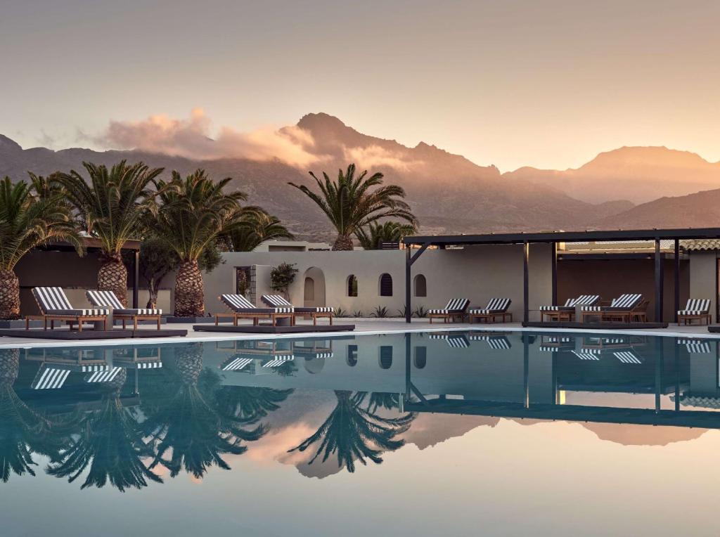 un hôtel avec une piscine bordée de palmiers et de montagnes dans l'établissement Numo Ierapetra Beach Resort Crete, Curio Collection Hilton, à Ierápetra