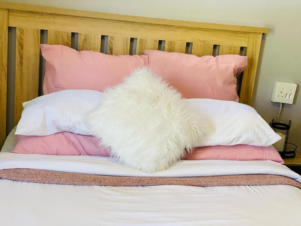 a white furry pillow on a bed with pink pillows at The M-Pire Cottage in Bloemfontein