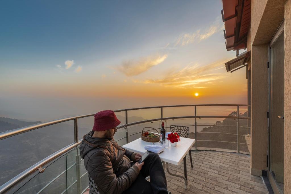 un hombre sentado en una mesa en un balcón viendo la puesta de sol en 3BDR Cottage Kasauli by Humble stay, en Kasauli