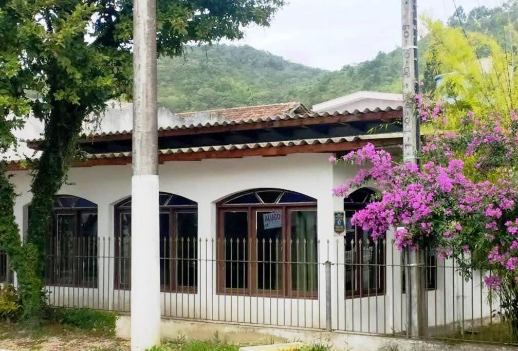 ein weißes Haus mit einem Zaun und lila Blumen in der Unterkunft Casa no centro da cidade a 600 metros da praia. in Porto Belo