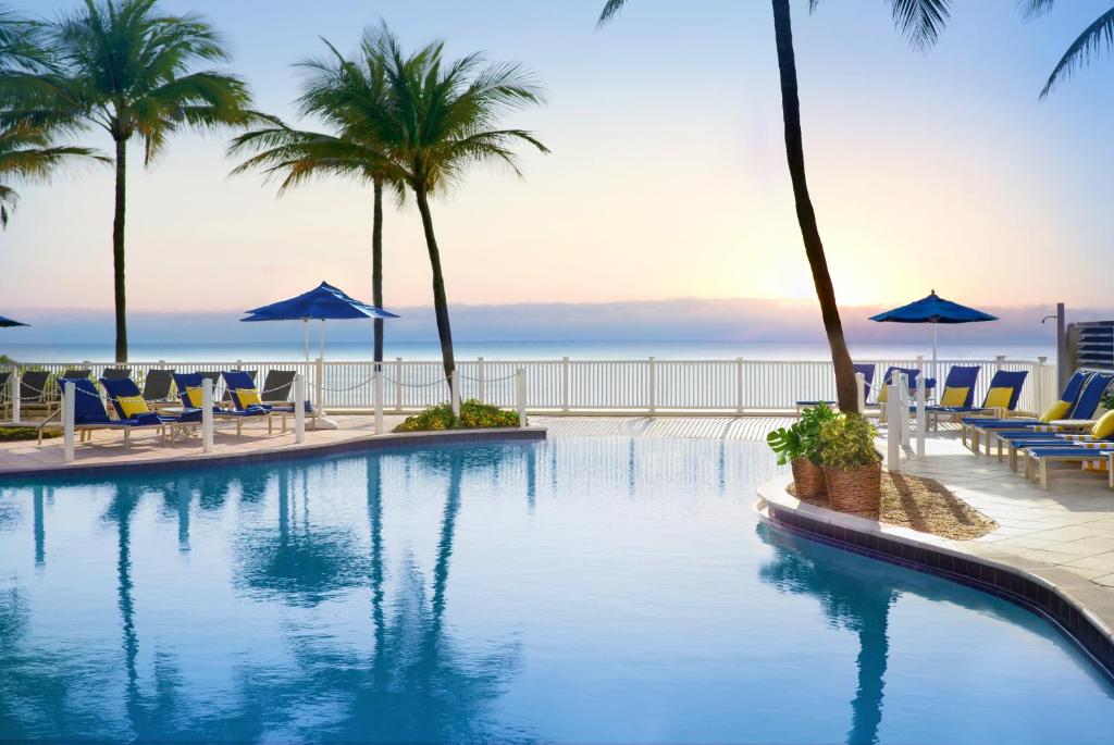 a swimming pool with chairs and palm trees and the ocean at Pelican Grand Beach Resort, a Noble House Resort in Fort Lauderdale