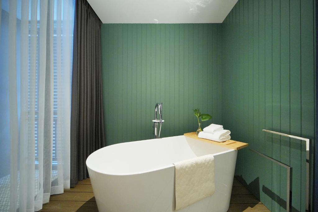 A freestanding white bathtub set against a green paneled wall beside a curtained window.