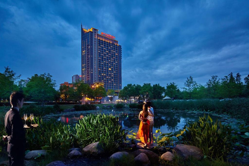 a couple standing in front of a pond with a building at Songjiang New Century Grand Hotel Shanghai in Songjiang