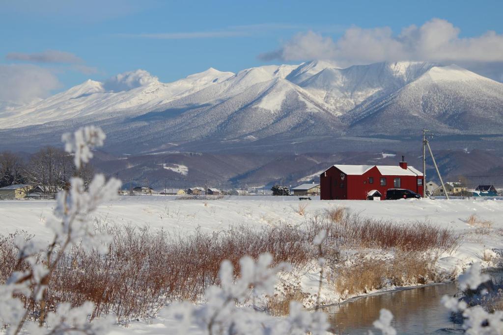 a red barn in a snowy field with mountains in the background at 縁 EN - The Suites in Furano
