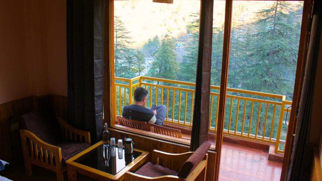 un uomo seduto su un balcone che guarda fuori da una finestra di The Woodpecker Inn Jibhi a Jibhi