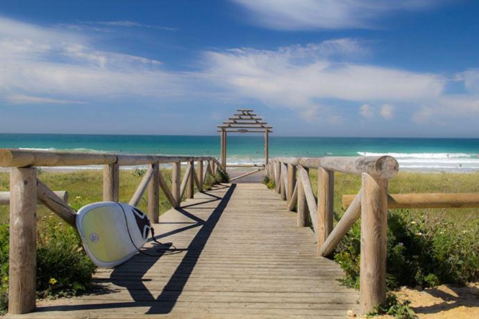 un paseo marítimo de madera que conduce a la playa con una tabla de surf en Coqueta casita de playa en el Palmar, en El Palmar