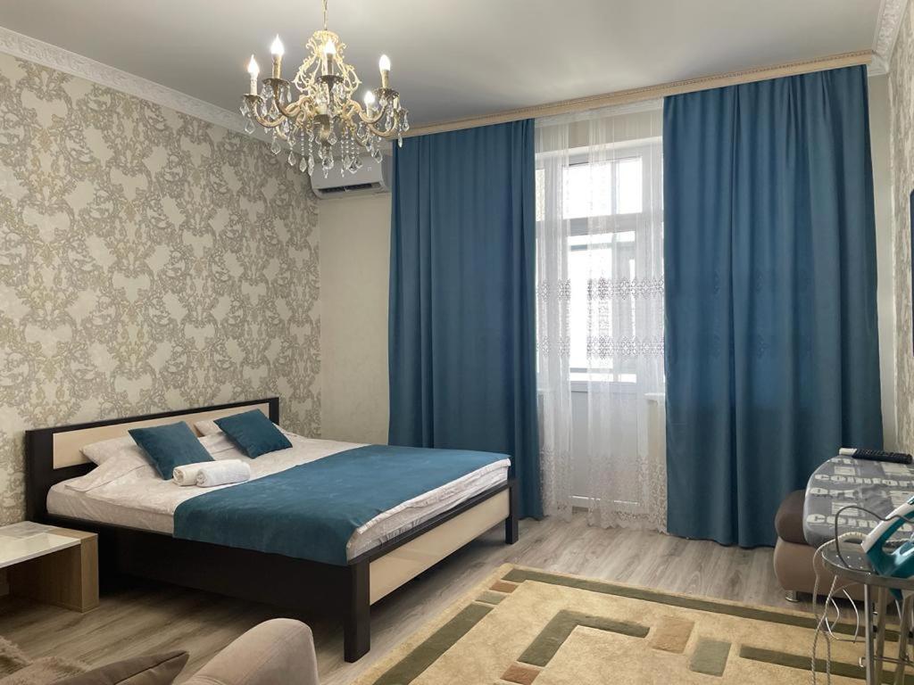 a bedroom with a bed with blue curtains and a chandelier at Apartment Mangilik El in Prigorodnyy