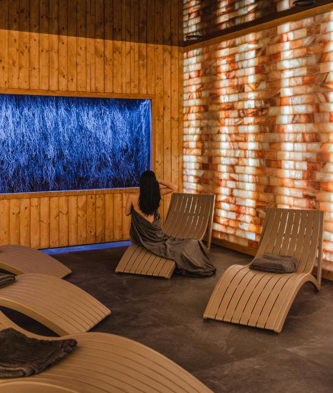 Woman wrapped in towel seated on wooden lounger in warm salt-wall relaxation room, calming ambient lighting.