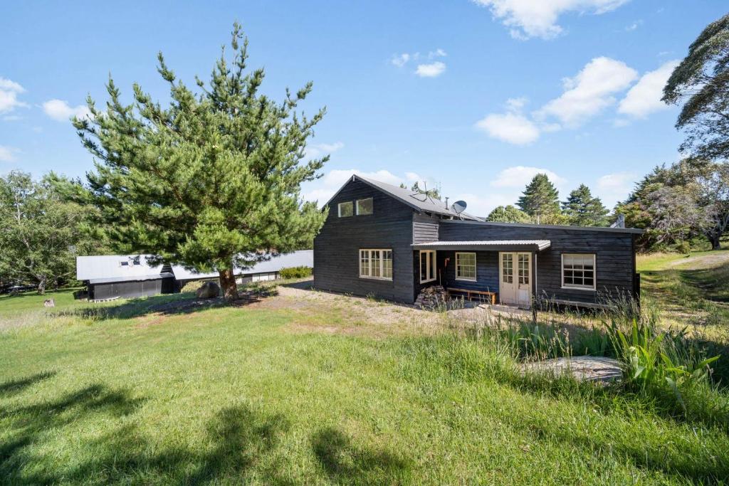 a black house in a field with a tree at Tukidale Cottage - 3BR Cottage l WiFi l Air-Con l Relaxing Location in Crackenback