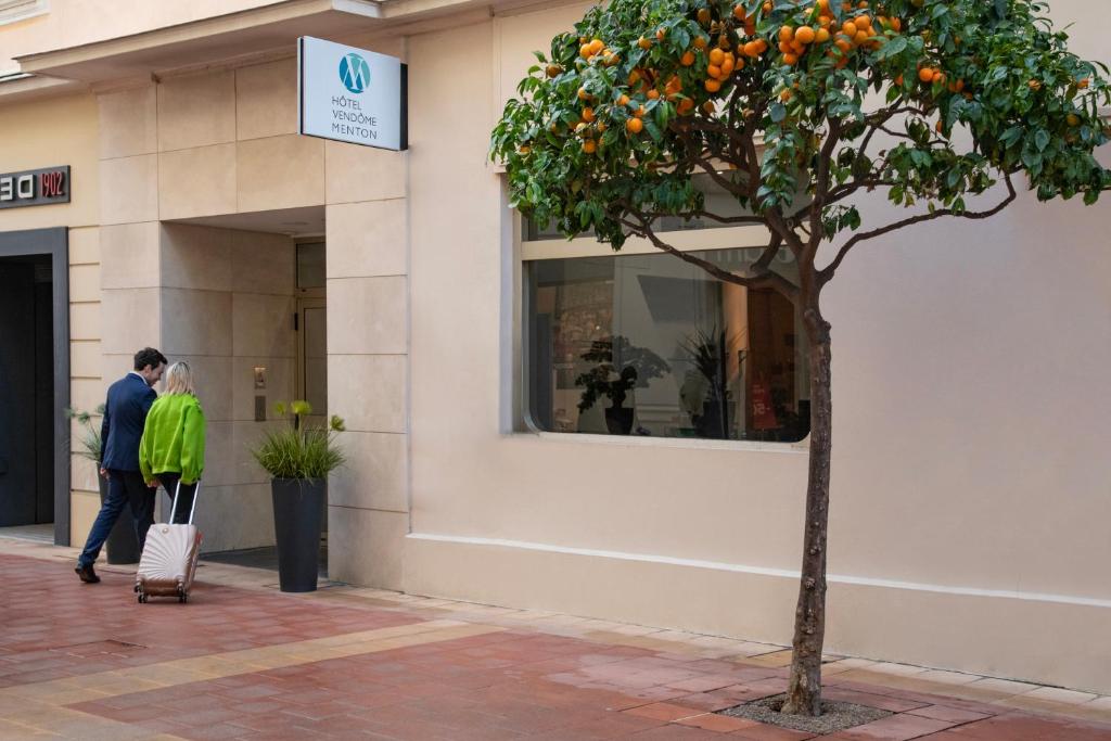 a man and woman walking in front of a building with an orange tree at Hôtel Vendôme Menton in Menton