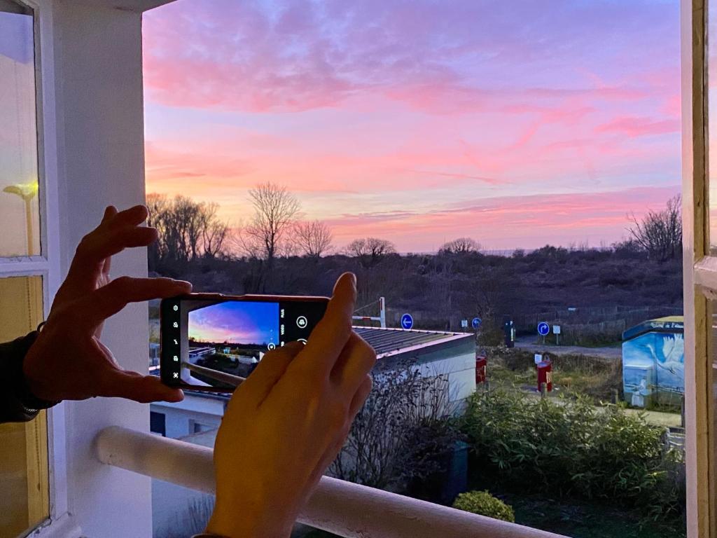 une personne prenant une photo d'un coucher de soleil depuis une fenêtre dans l'établissement Le Gîte du Phare vue sur mer, à Cayeux-sur-Mer