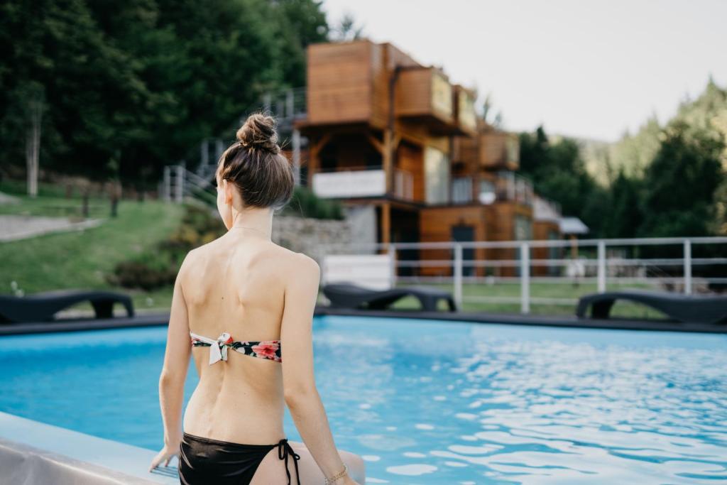 Young woman in a bikini at the pool edge, looking toward stacked wooden cabins and surrounding greenery.