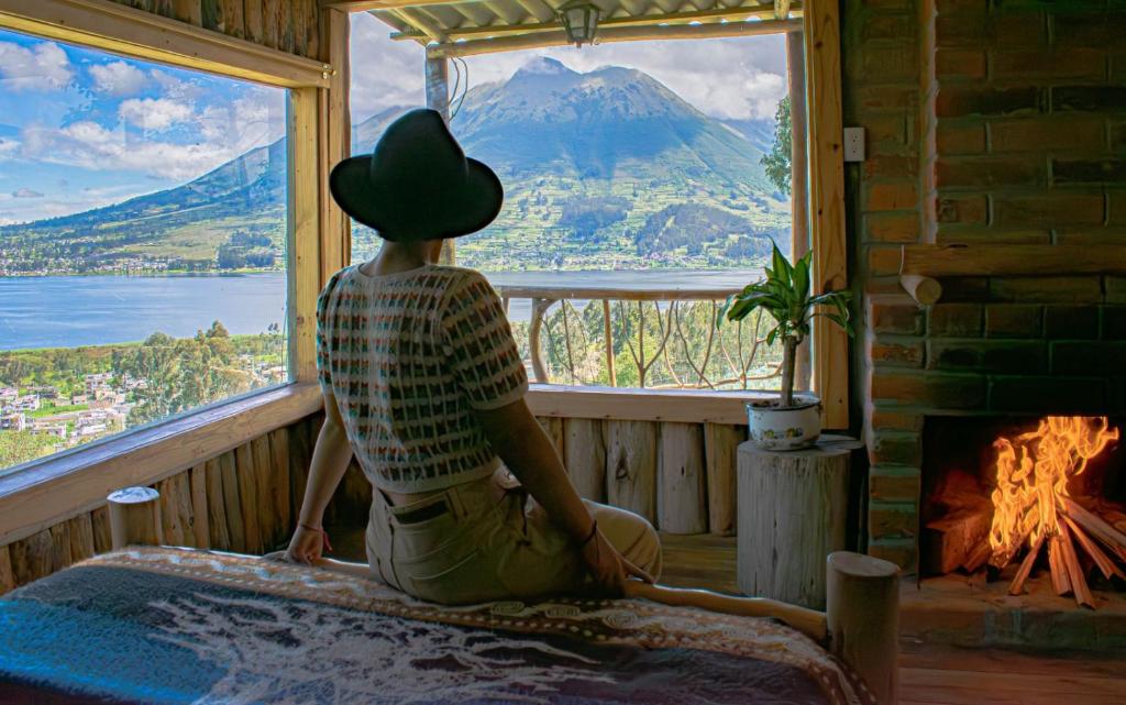 osoba w kapeluszu siedząca na łóżku patrząca przez okno w obiekcie Cabañas y Glampings Balcon del lago w mieście Otavalo