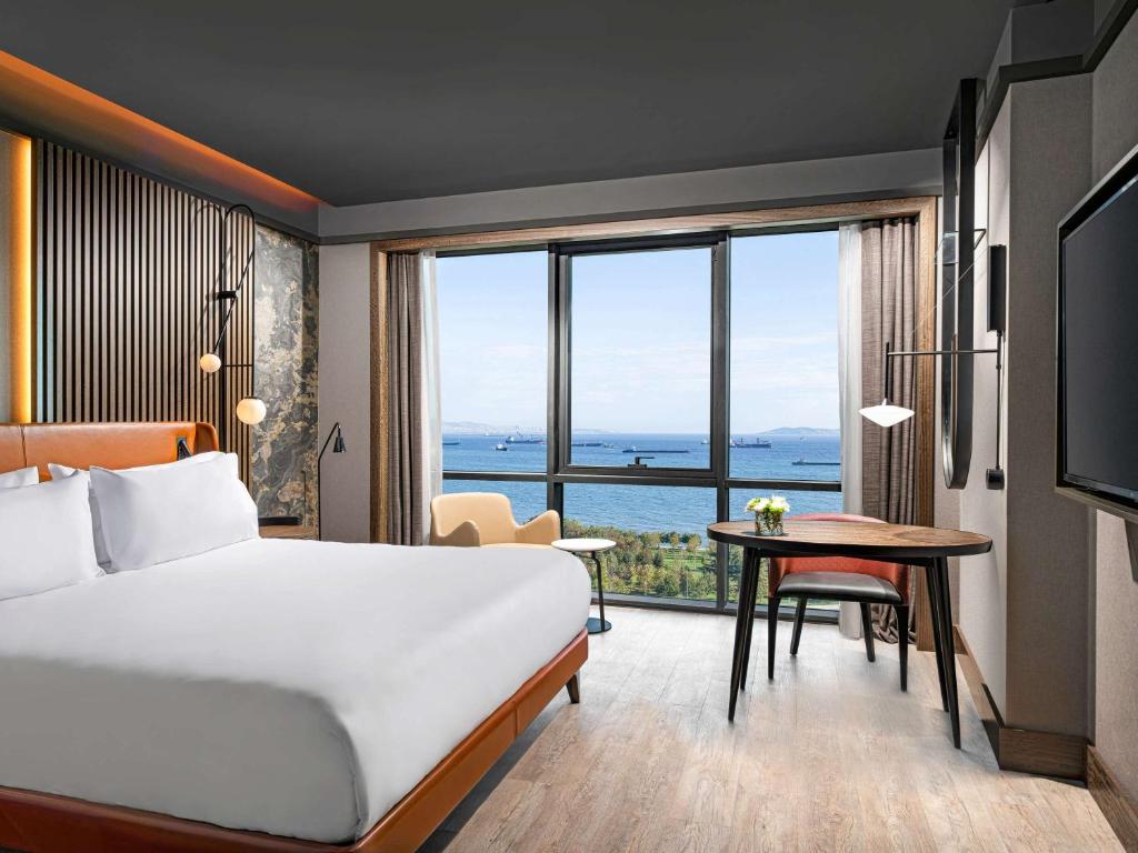 イスタンブールにあるMövenpick Hotel Istanbul Marmara Seaのベッド、デスク、大きな窓が備わるホテルルームです。