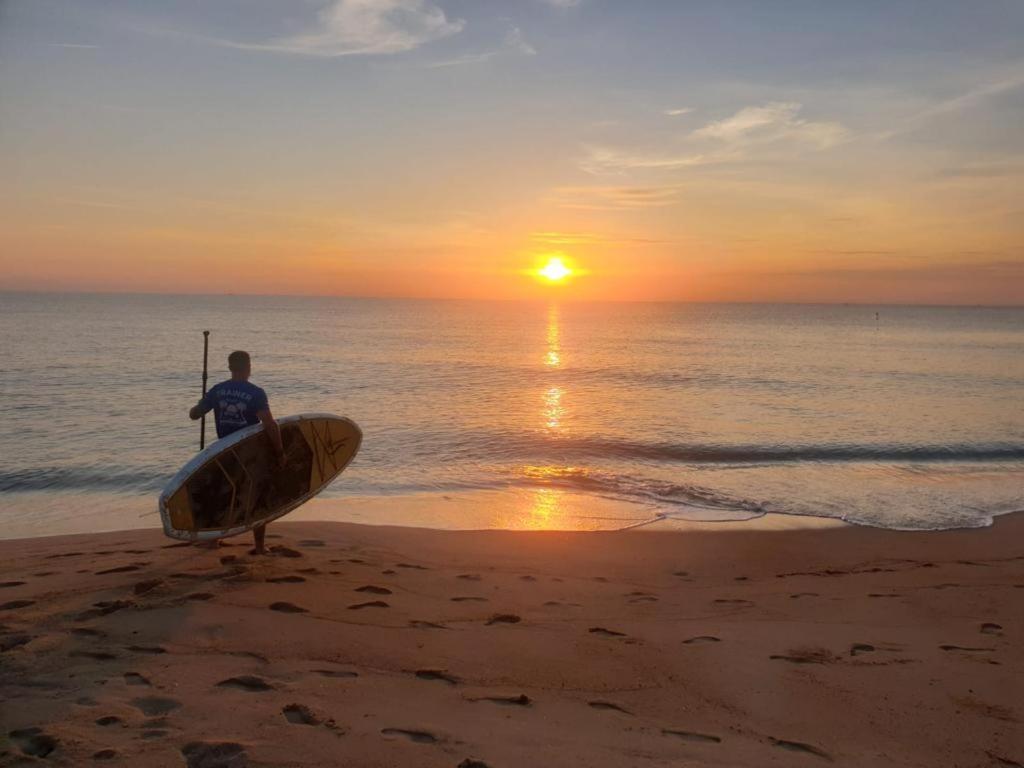 Ein Mann steht am Strand mit einem Surfbrett in der Unterkunft Loma Beach Resort in Ban Pak Nam Pak Duat