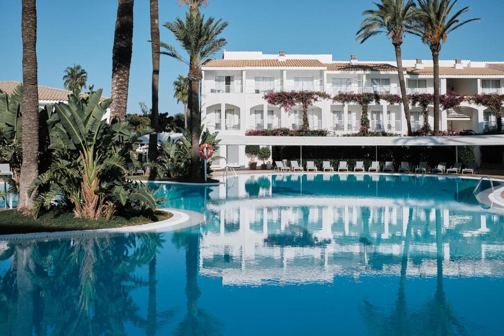 una piscina con palmeras y un edificio en Prinsotel La Caleta en Ciutadella