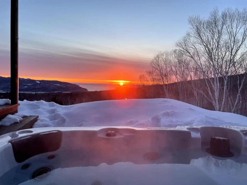 un bain à remous recouvert de neige avec le coucher du soleil en arrière-plan dans l'établissement Cozy Haven with Spa & Stunning Views, à Baie-Saint-Paul