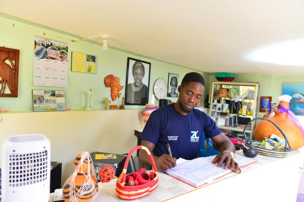 un homme assis à un comptoir écrivant sur un papier dans l'établissement Zion Camp, à Entebbe