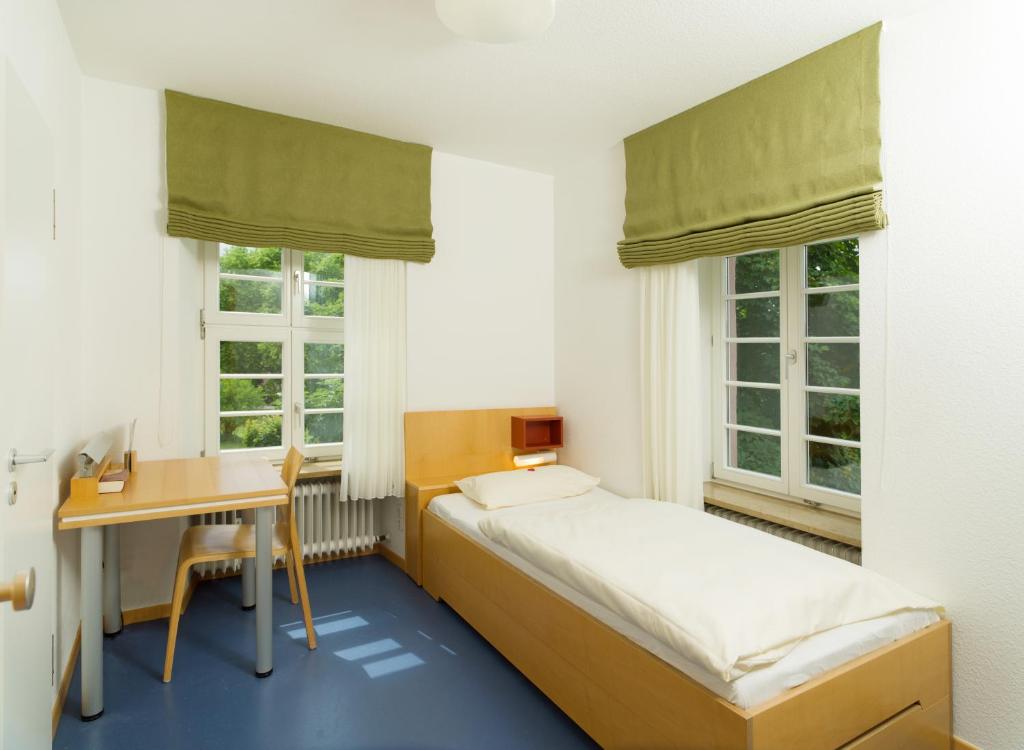 a bedroom with a bed and a desk and two windows at Bildungshaus Neckarelz in Mosbach